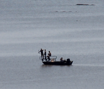 [Several people on an open top motorboat with rails at the front. One person has a bow outstretched pointing down toward the water in front of the boat.]